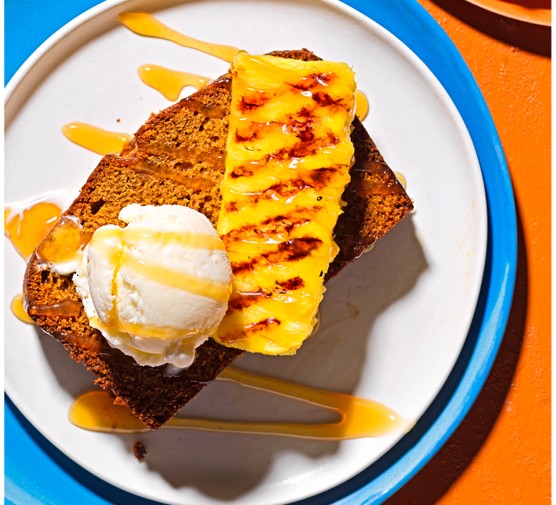 Rum cake recipe A slice of ginger and rum cake topped with a scoop of ice cream and a wedge of pineapple, on a blue plate with an orange background