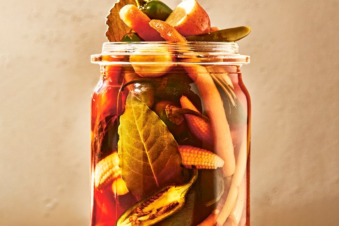 Chiles en escabeche a glass jar on a stone surface overflowing with chillies, bay leaves, sliced potatoes and baby corn