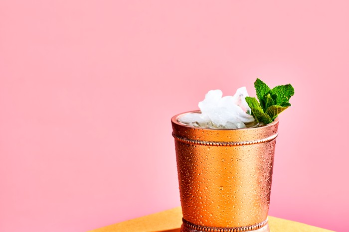 mint julep A copper cup filled with crushed ice and a sprig of mint on a yellow table with a pink background