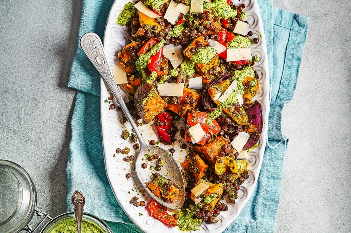 Roast vegetable salad with kale pesto A grey background with a white platter, topped with roasted vegetables, grains and a green dressing