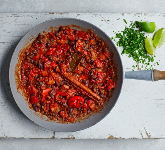 Caribbean-style black bean curry a silver pan with a wooden handle on a white wooden chopping board filled with pan of a bright-red coloured curry