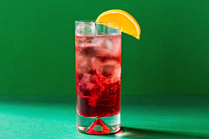 A green background with a tall glass filled with a red cocktail and a wedge of orange