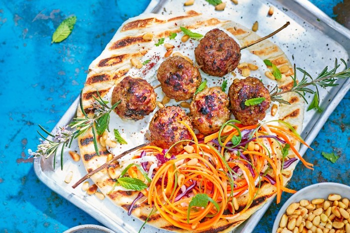 BBQ kofte with whipped feta and tahini slaw A silver tray topped with a large grilled flatbread meatballs and slaw on a blue background
