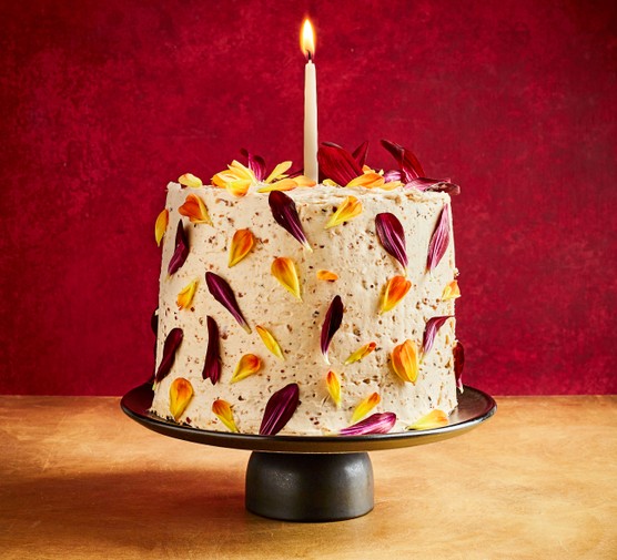 Celebration Cake Recipe A birthday cake covered in purple, yellow and orange petals on a cakestand with a candle on the top