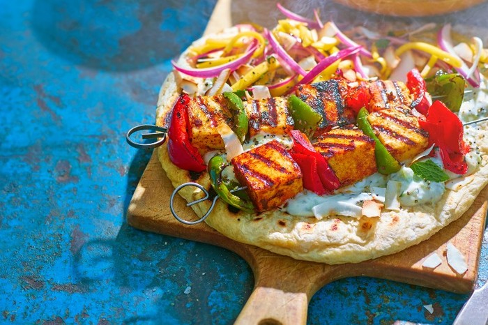 Chilli paneer skewers A round flatbread topped with bbq paneer skewers on a wooden board with a blue background