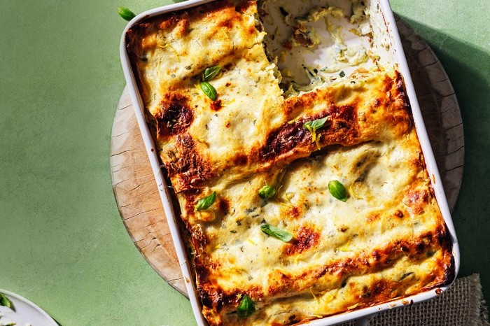 Courgette, parma ham and lemon ricotta lasagne A white ceramic oven dish filled with baked lasagne on a wooden board and a green surface