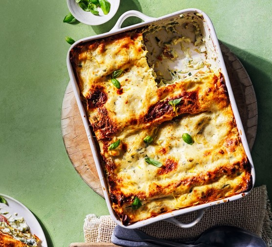 Courgette, parma ham and lemon ricotta lasagne A white ceramic oven dish filled with baked lasagne on a wooden board and a green surface
