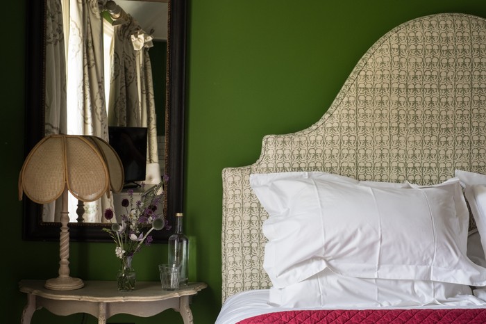 Room Five at The Swan Oxfordshire A bedroom with pea green walls, a cream fabric head board and a side table with flowers on