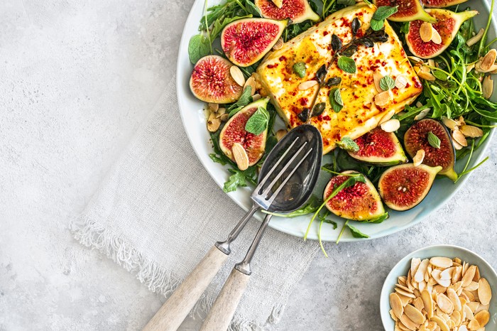 Baked feta salad with figs and almonds A large white serving plate filled with green leaves and topped with halved figs and a block of baked feta, on a light stone background
