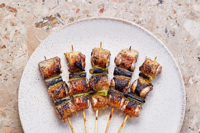 Humble Chicken Chicken thigh yakitori A white mottled plate on a stone background with five bamboo skewers fanned out, threaded with charred chicken