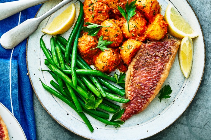 Red mullet with romesco potato salad A grey plate topped with pan-fried fish, golden potatoes, green beans and slices of yellow lemons