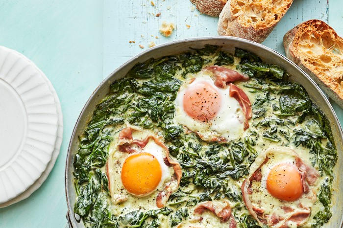 Florentine Spinach Packed Eggs Metal Pan Filled with Creamy Spinach and Eggs Next to Slices of Ciabatta on a Blue Background