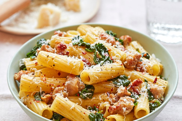 Sausage, spinach and chilli pasta A bowl of rigatoni pasta with spinach and meat, with a plate of parmesan in the background on a white cloth