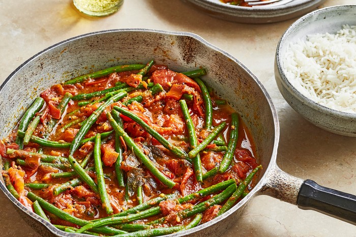 Easy Green Bean Curry Recipe a metal pan containing green beans and tomato with a bowl of rice on a stone background