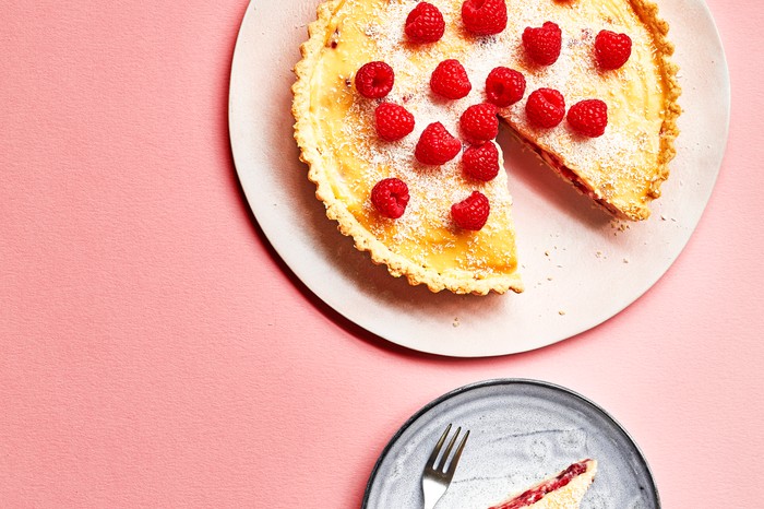 A custard tart topped with raspberries and dusted with icing sugar on a white plate with a pink background