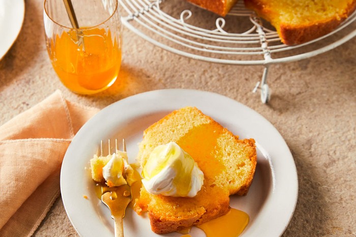 Orange Drizzle Cake Recipe An orange drizzle cake loaf on a cake stand with a slice of cake on a separate plate topped with a dollop of cream