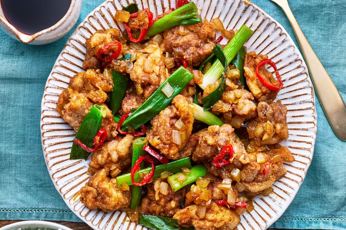 Salt and Pepper Chicken Recipe A plate of chicken with spring onions on a turquoise tablecloth with a bowl of rice and a spoon on the side