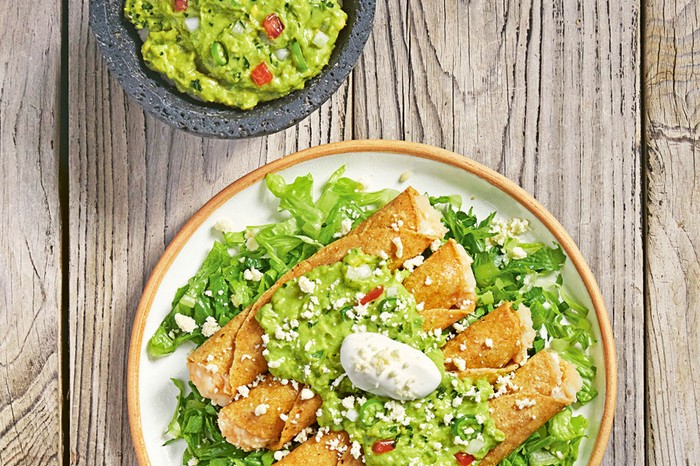 Taquitos dorados de papa Rolled potato tacos on a bed of salad topped with guacamole and sour cream, next to a bowl of guacamole